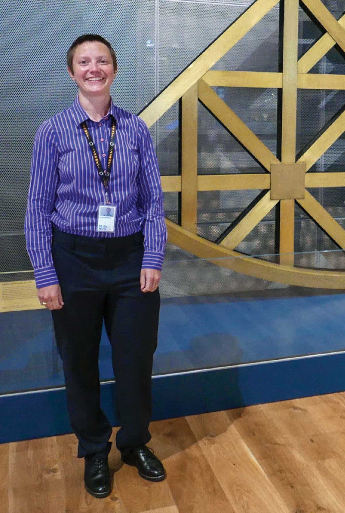 Smiling young woman with short brown hair wearing a striped purple, tucked-in shirt and black pants and shoes standing in front of a large scientific instrument