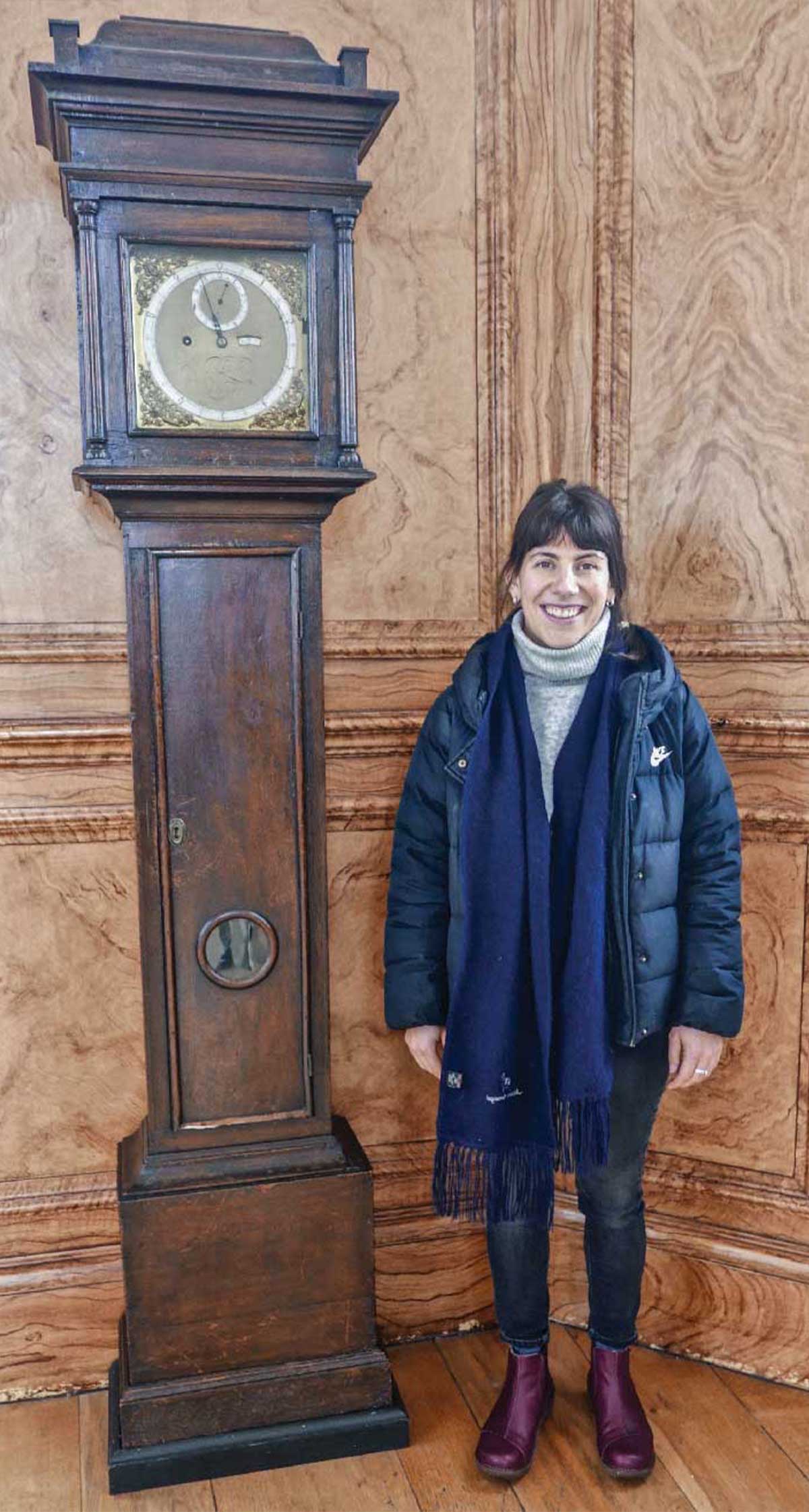 Smiling woman with black hair wearing a blue jacket and scarf standing beside a tall case clock
