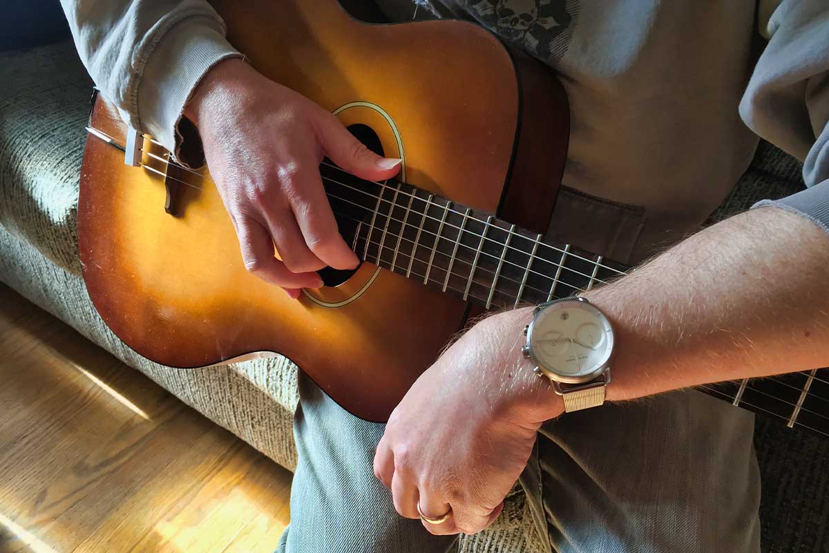 A man sitting on a couch holding an acoustic guitar wearing a Nordgreen Pioneer chronograph on his wrist